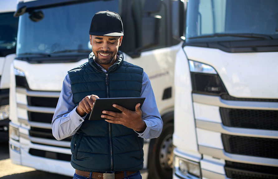 man in vest and hat using tablet in front of two parked trucks